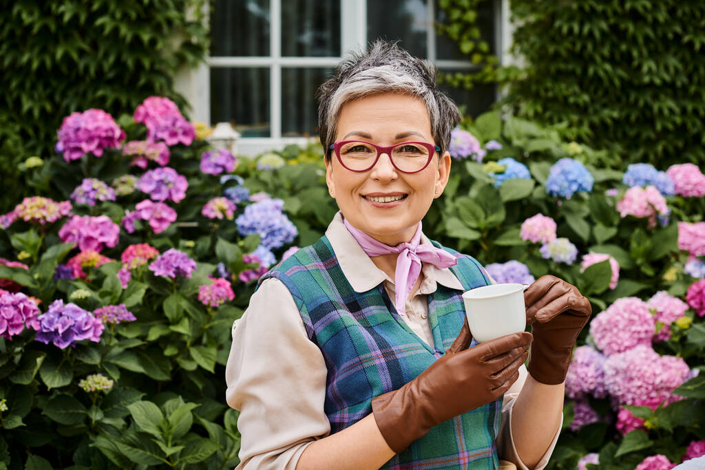 elegant jolly mature woman in chic attire drinking tea near house in England and smiling at camera - Photo, Image