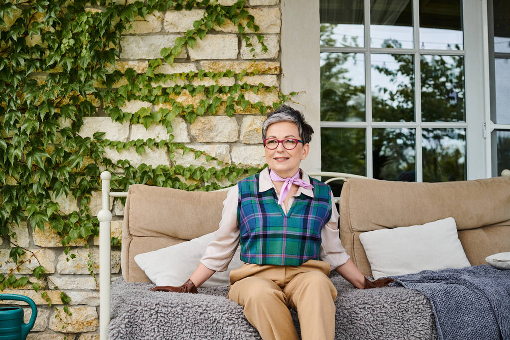 ελκυστική χαρούμενη ώριμη γυναίκα κάθεται στον καναπέ κοντά στο σπίτι της στην Αγγλία και κοιτάζοντας την κάμερα - Φωτογραφία, εικόνα