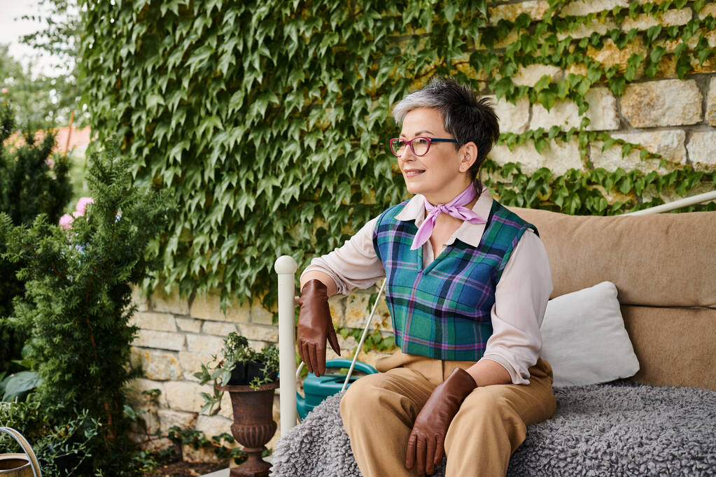 goed uitziende vrolijke volwassen vrouw zitten op bank in de buurt van haar huis in Engeland en kijken weg - Foto, afbeelding
