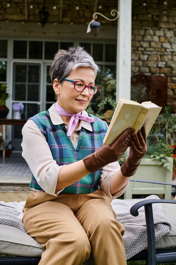витончена зріла радісна жінка в окулярах читає книгу біля свого будинку в сільській Англії - Фото, зображення
