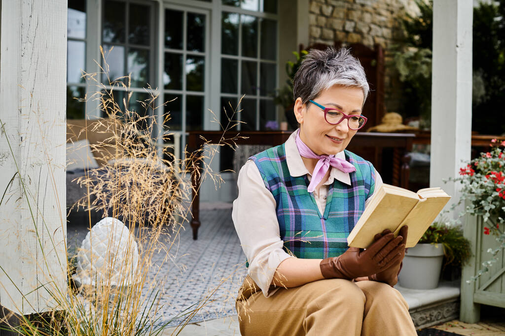 kifinomult érett vidám nő szemüveges olvasókönyv közelében háza vidéki Angliában - Fotó, kép