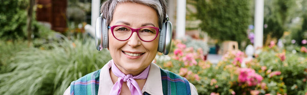 debonair femme mûre dans des lunettes élégantes posant avec des écouteurs et souriant à la caméra, bannière - Photo, image