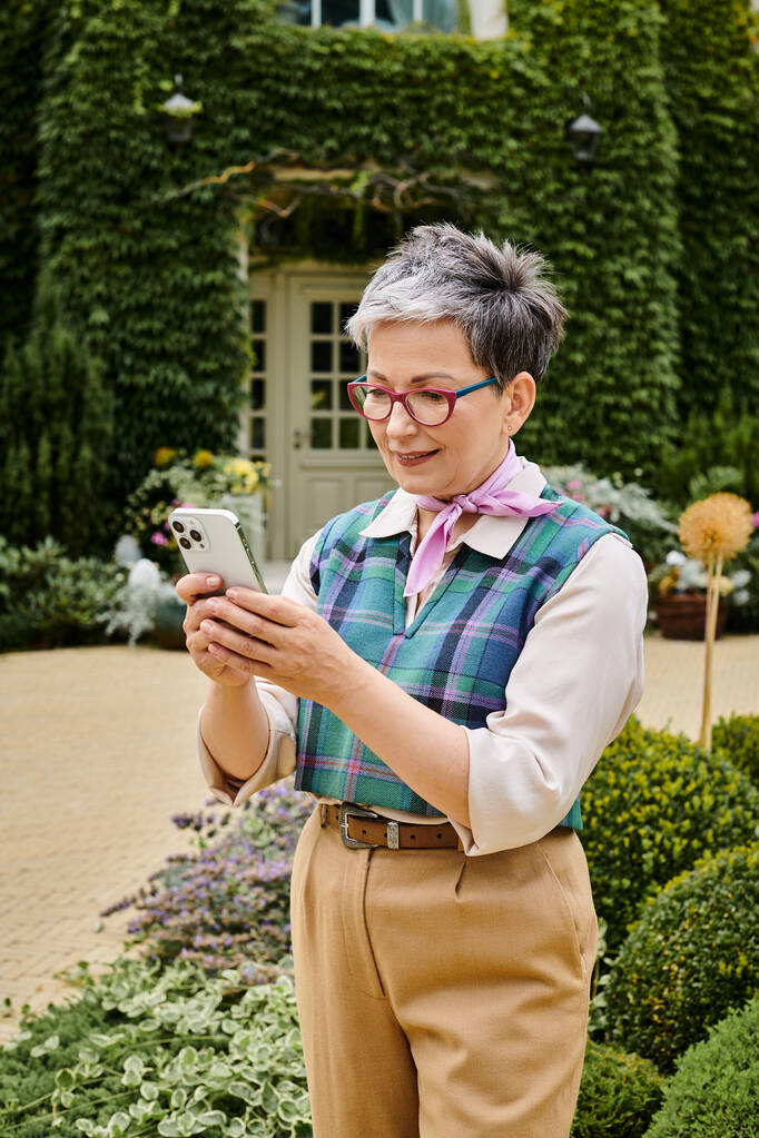 витончена щаслива зріла жінка в шикарному вбранні, дивлячись на її смартфон біля її будинку в Англії - Фото, зображення