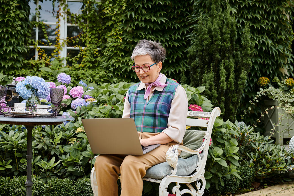 debonair ώριμη εύθυμη γυναίκα με σικ ενδυμασία κάθεται με φορητό υπολογιστή κατά την ώρα του τσαγιού κοντά στο σπίτι της στην Αγγλία - Φωτογραφία, εικόνα