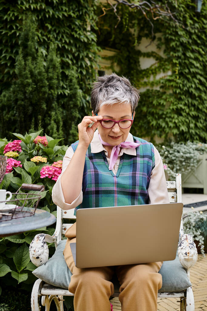 wyrafinowany dojrzała wesoła kobieta w eleganckim stroju siedzi z laptopem w czasie herbaty w pobliżu jej domu w Anglii - Zdjęcie, obraz