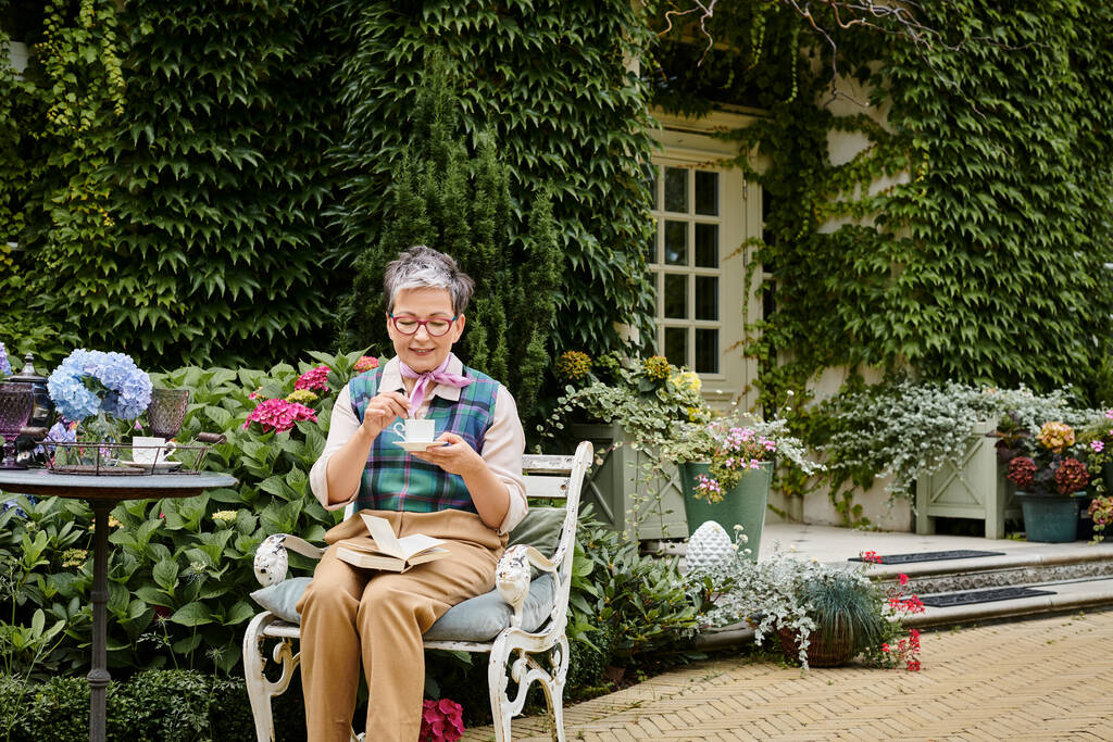 εκλεπτυσμένη ώριμη χαρούμενη γυναίκα με γυαλιά απολαμβάνοντας ζεστό τσάι και το βιβλίο κοντά στο σπίτι της στην Αγγλία - Φωτογραφία, εικόνα