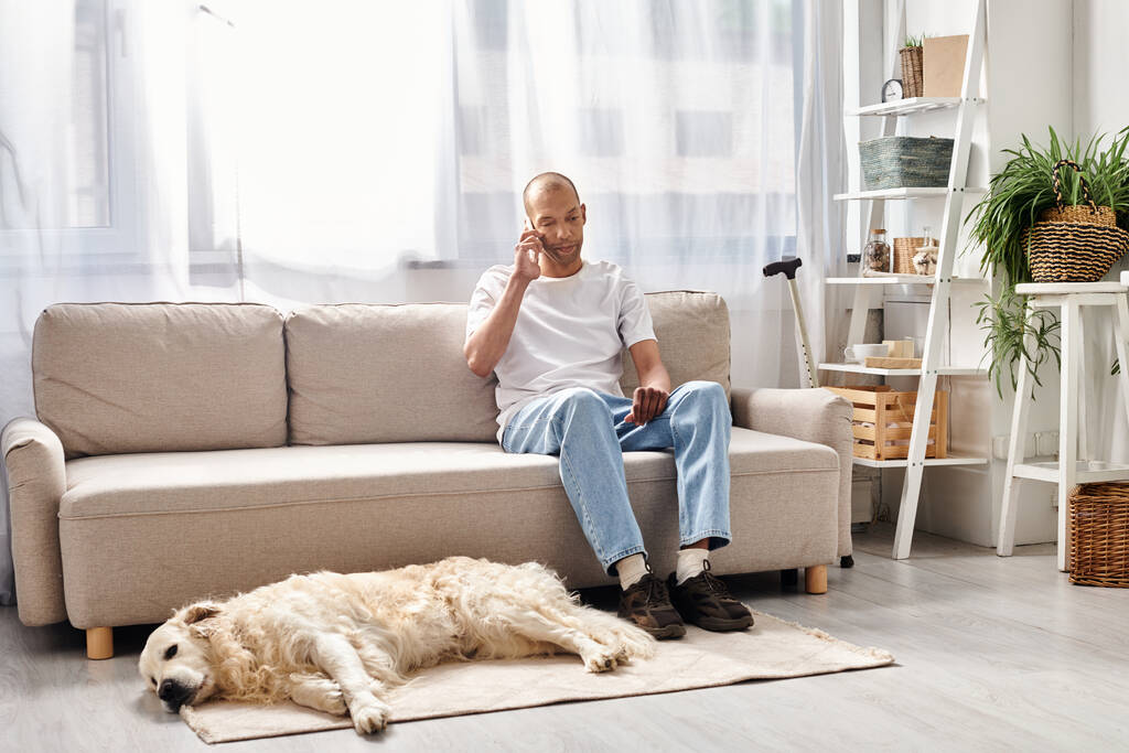 Афроамериканец с миастенией Гравис сидит на диване в сопровождении своей верной лабрадорской собаки в уютной обстановке.. - Фото, изображение