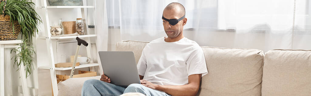 Un hombre afroamericano con síndrome de Miastenia Gravis está sentado en un sofá, usando una computadora portátil, con su perro Labrador cerca. - Foto, Imagen