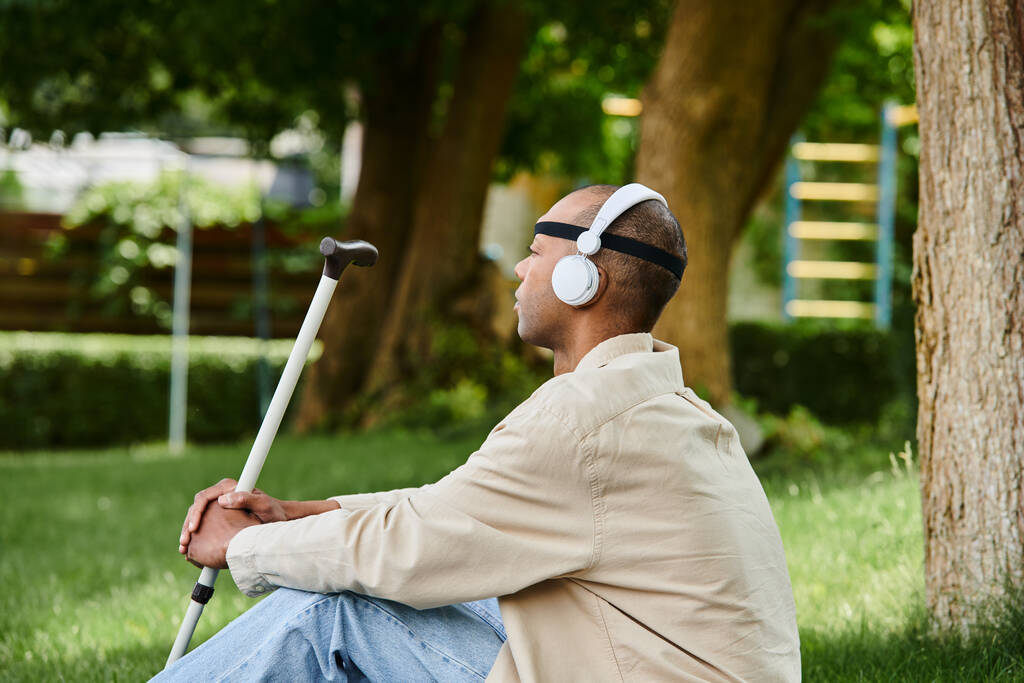 Ένας Αφροαμερικάνος με σύνδρομο μυασθένεια gravis κάθεται στο γρασίδι με ακουστικά, απολαμβάνοντας μουσική. - Φωτογραφία, εικόνα