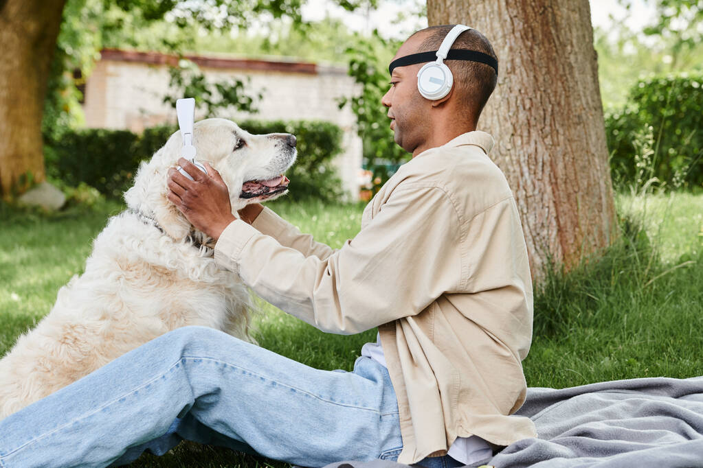 Ένας ανάπηρος Αφροαμερικανός με σύνδρομο μυασθένεια Gravis κάθεται στο γρασίδι με ένα σκύλο Λαμπραντόρ που φοράει ακουστικά, απολαμβάνοντας μια ειρηνική στιγμή μαζί. - Φωτογραφία, εικόνα