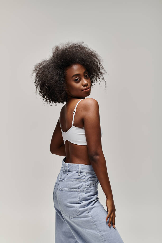 Красивая молодая афроамериканка с вьющимися волосами в белом топе и голубыми брюками, излучающими элегантность и изящество. - Фото, изображение