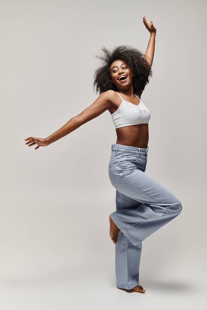 Güzel, kıvırcık saçlı, genç bir Afrikalı Amerikalı kadın stüdyo ortamında beyaz bir bluzla enerjik bir şekilde dans ediyor.. - Fotoğraf, Görsel