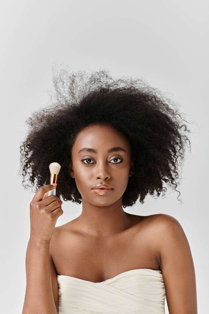 Joven mujer afroamericana con cabello rizado sosteniendo cepillo cosmético en un entorno de estudio que representa el concepto de belleza. - Foto, imagen