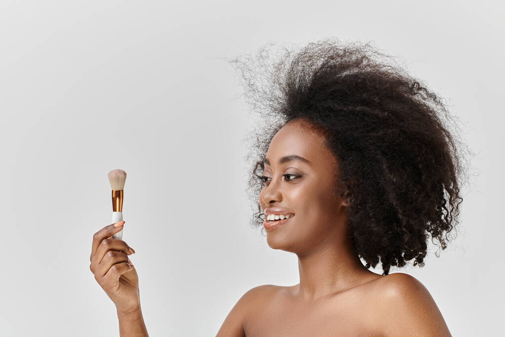 Mujer afroamericana desnuda con cabello rizado sosteniendo cepillo cosmético, exudando belleza y confianza en un entorno de estudio. - Foto, Imagen