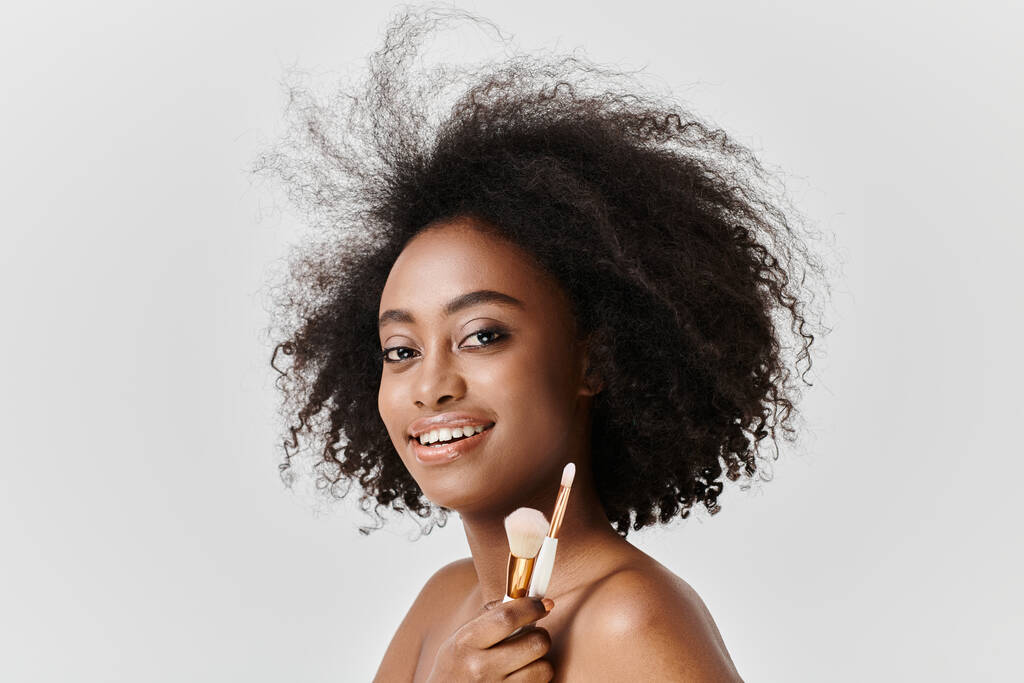 Mulher afro-americana com cabelo encaracolado segurando pincéis de maquiagem, aplicando maquiagem em um estúdio. - Foto, Imagem