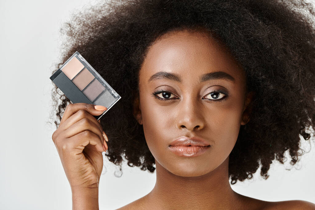 Eine schöne junge Afroamerikanerin mit lockigem Haar hält eine Palette an Make-up vor ihrem Gesicht. - Foto, Bild