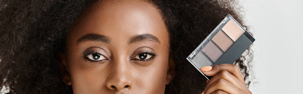 Žena s kudrnatými vlasy drží make-up paletu před obličejem, předvádí umění a krásu ve studiu nastavení. - Fotografie, Obrázek