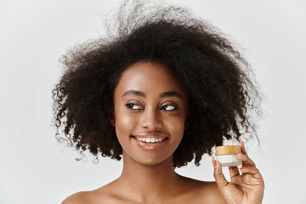 Una hermosa joven afroamericana con el pelo rizado sostiene un frasco de crema, encarnando la esencia del cuidado de la piel. - Foto, Imagen