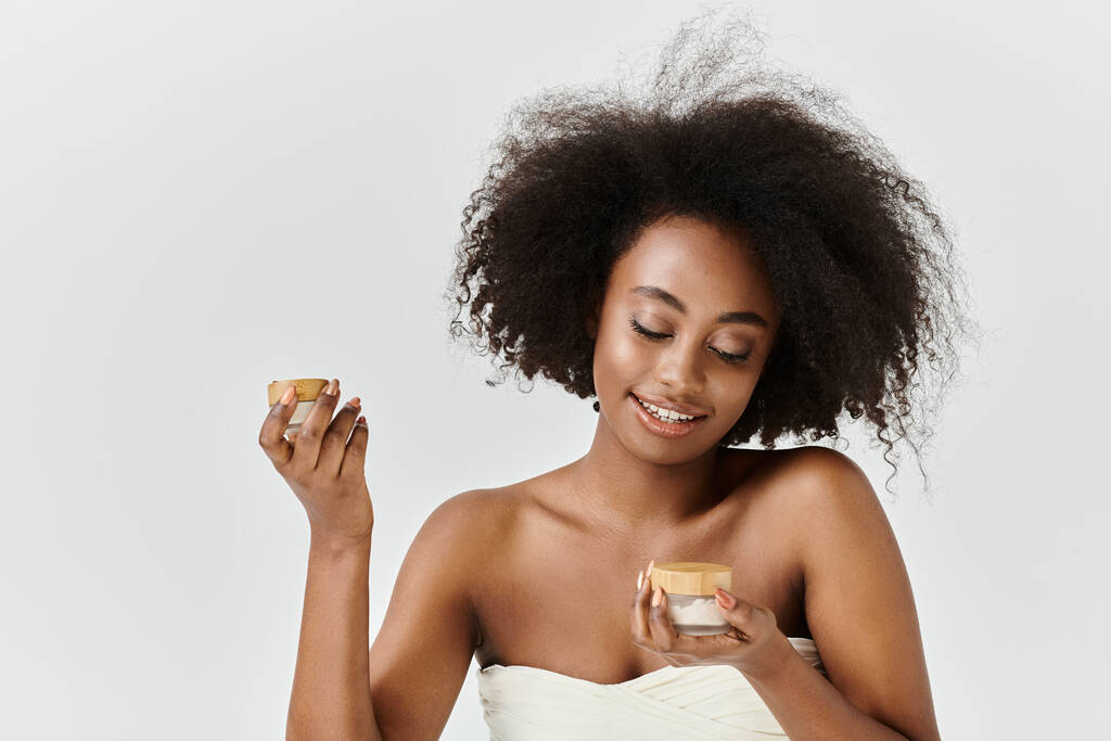 Una splendida giovane donna afroamericana, avvolta in un asciugamano, tiene un barattolo con crema - Foto, immagini