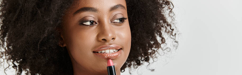 Piękna, młoda Afroamerykanka z kręconymi włosami elegancko trzyma szminkę w studio - Zdjęcie, obraz