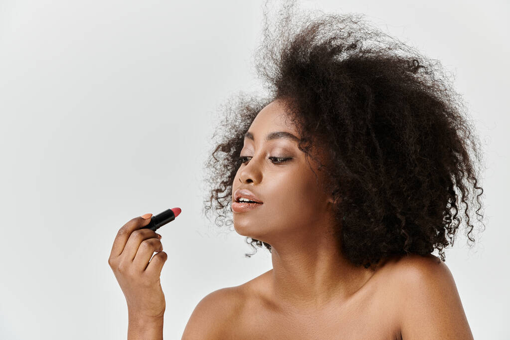 A beautiful young African American woman with curly hair applying lipstick, in a studio setting - Photo, Image
