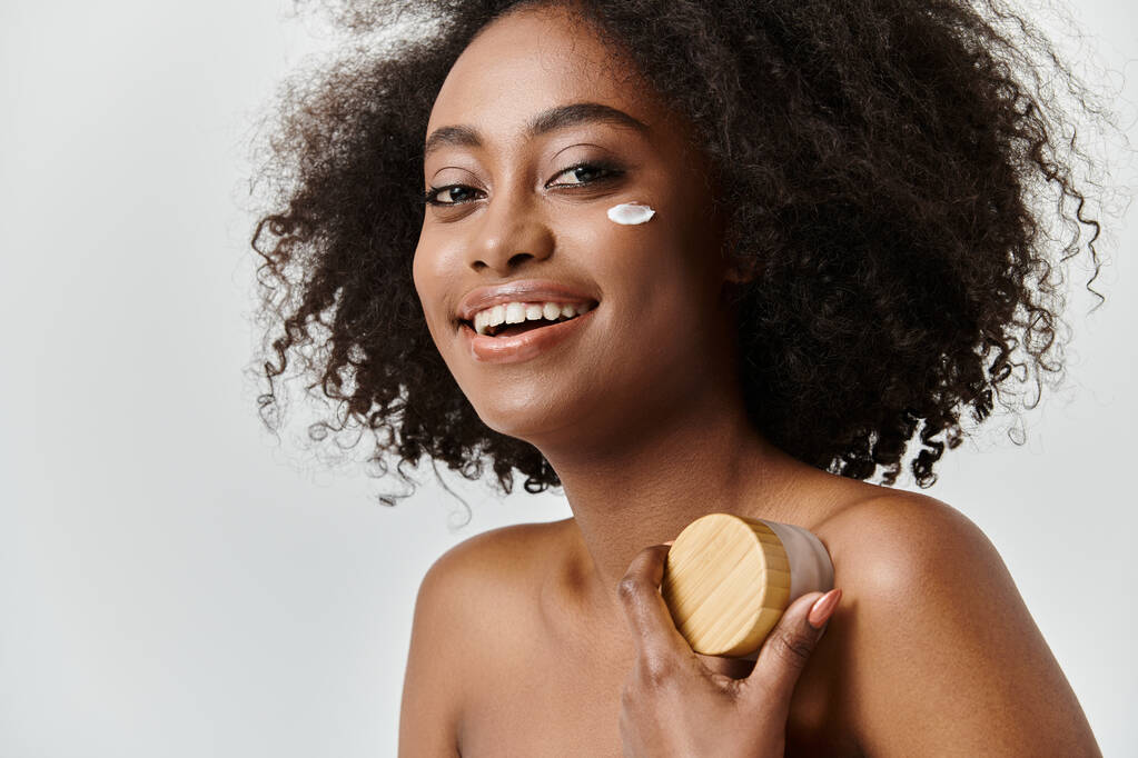 Una bella giovane donna afroamericana con i capelli ricci tiene una crema con copertura in legno in un ambiente di studio, trasudando un concetto di cura della pelle. - Foto, immagini