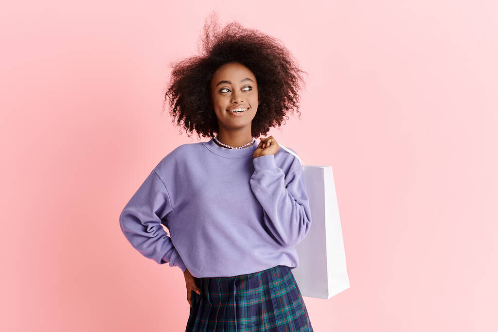 Красивая молодая афроамериканка с вьющимися волосами в фиолетовом свитере и клетчатой юбкой, держащая сумку с покупками.. - Фото, изображение