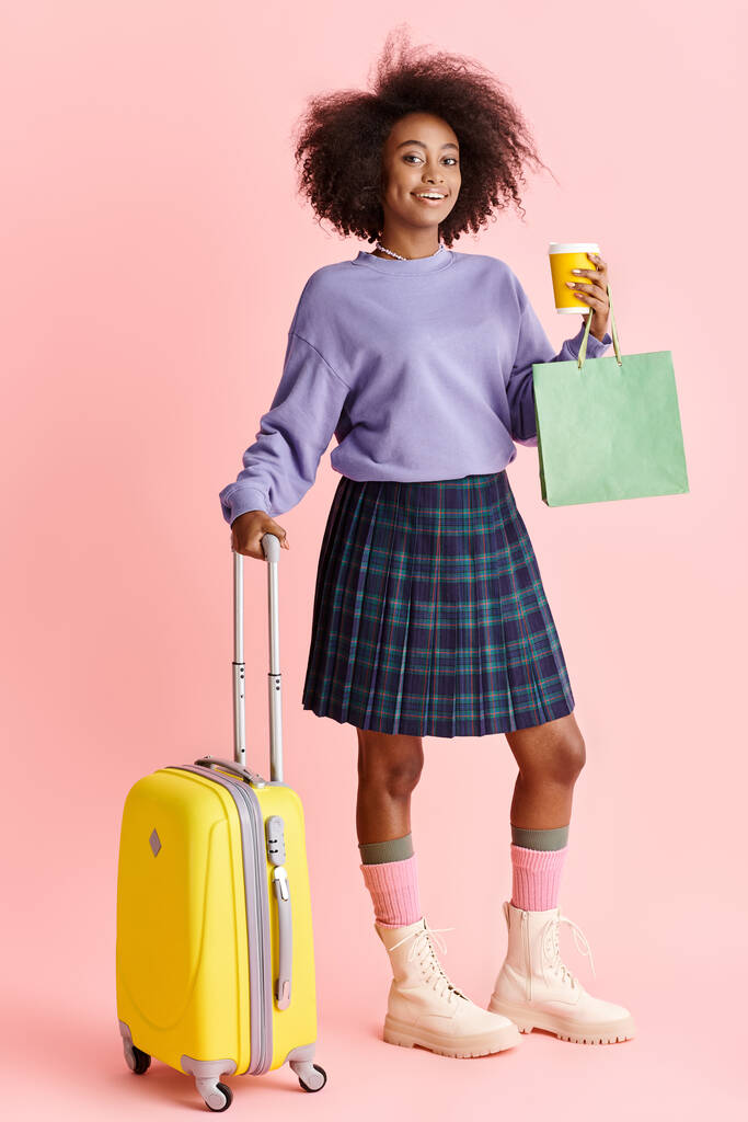 Giovane donna afroamericana in maglione viola e gonna a quadri, pronta per viaggiare con una valigia gialla brillante. - Foto, immagini