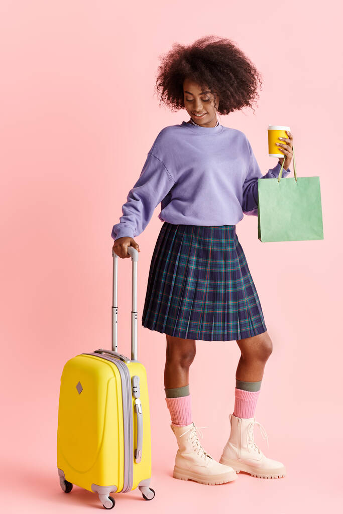 Kaunis nuori afroamerikkalainen nainen, jolla on kiharat hiukset violetissa paidassa ja ruudullisessa hameessa, mukanaan keltainen matkalaukku.. - Valokuva, kuva