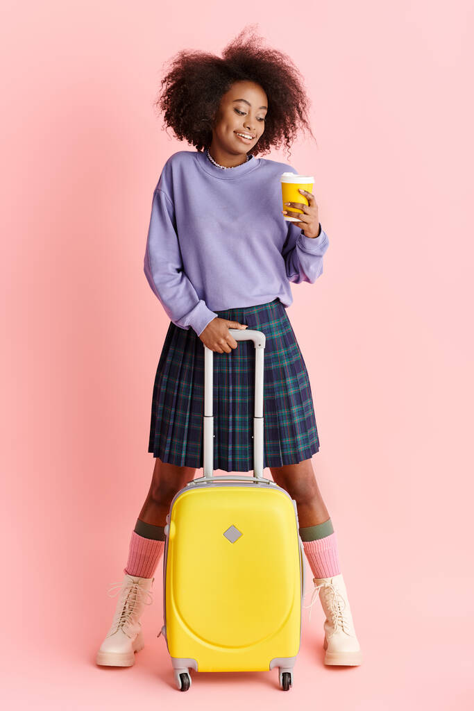 Mladá Afroameričanka v purpurovém svetru a kostkované sukni, držící žlutý kufr ve studiu. - Fotografie, Obrázek