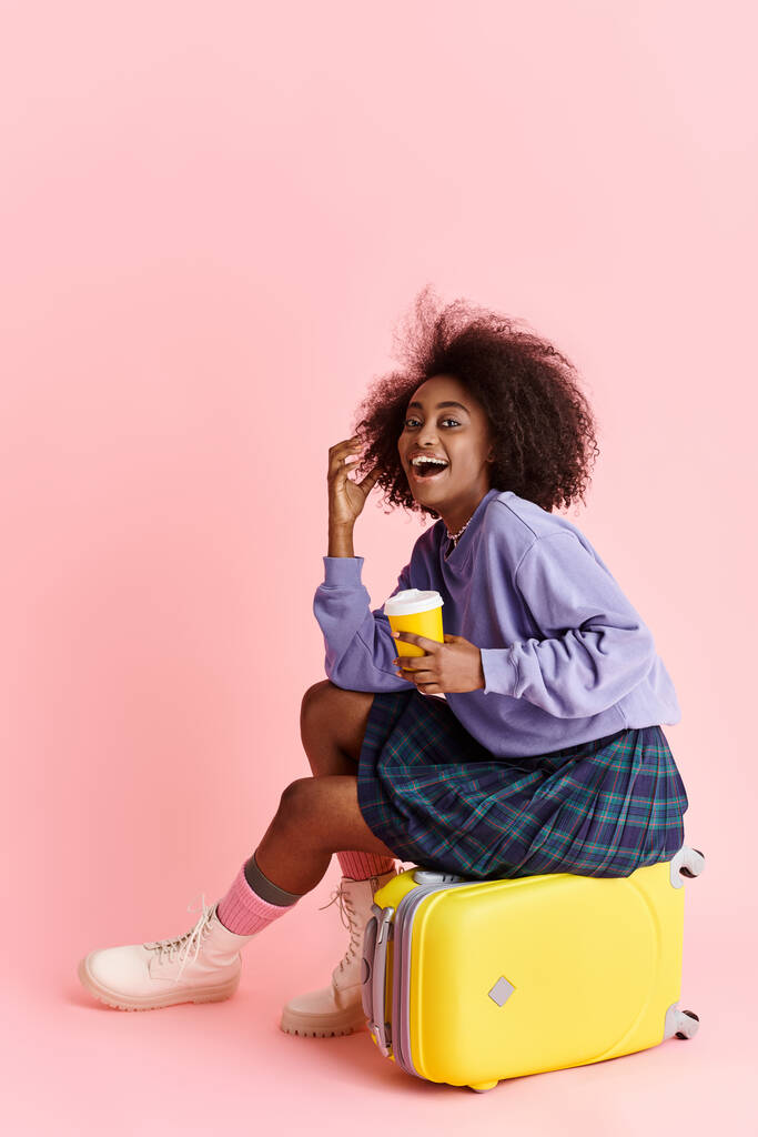 Стильна молода афроамериканка з кучерявим волоссям відпочиває на валізі, смакуючи чашку кави. - Фото, зображення