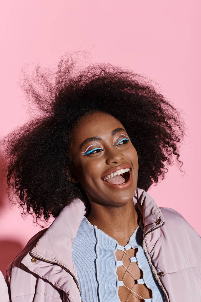 Een stijlvolle jonge Afro-Amerikaanse vrouw met krullend haar glimlacht helder in een studio omgeving, uitstralen van vertrouwen en vreugde. - Foto, afbeelding