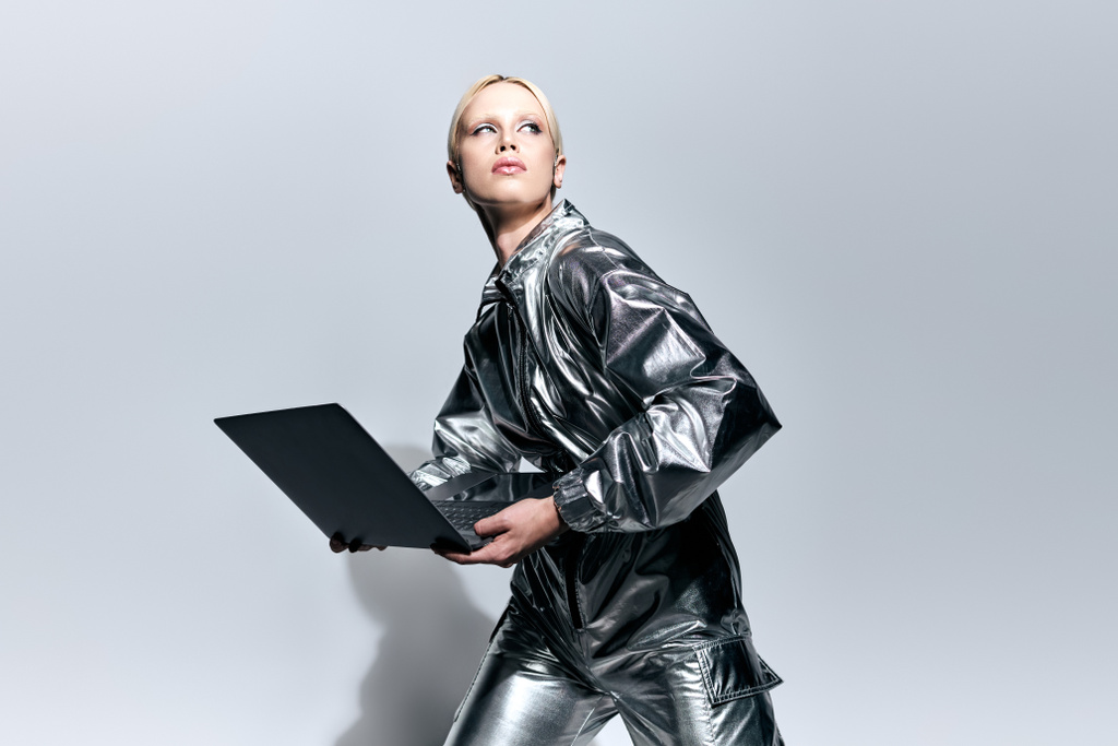 extravagantní krásná žena ve stříbrném robotickém oblečení pózuje v pohybu s notebookem a dívá se jinam - Fotografie, Obrázek