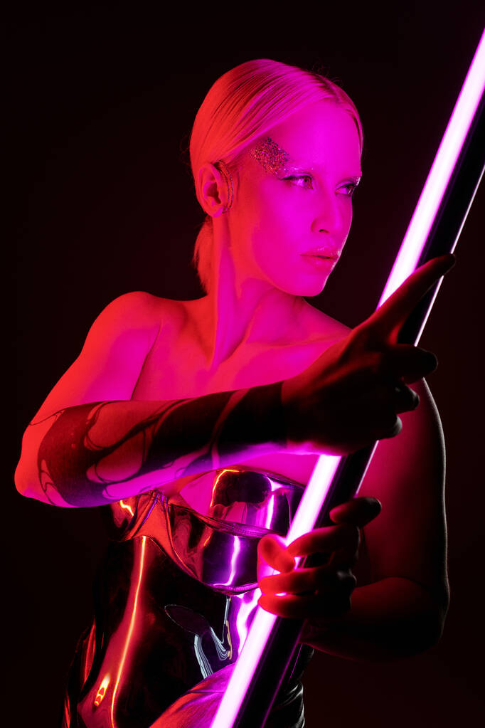 attraktive Frau in futuristischer metallischer Kleidung, die einen rosafarbenen LED-Lampenstab hält und wegschaut - Foto, Bild