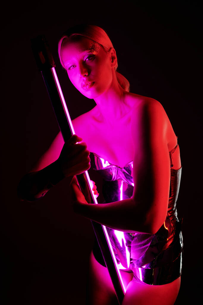 atractiva mujer en traje futurista metálico sosteniendo palo de lámpara led rosa y mirando a la cámara - Foto, Imagen