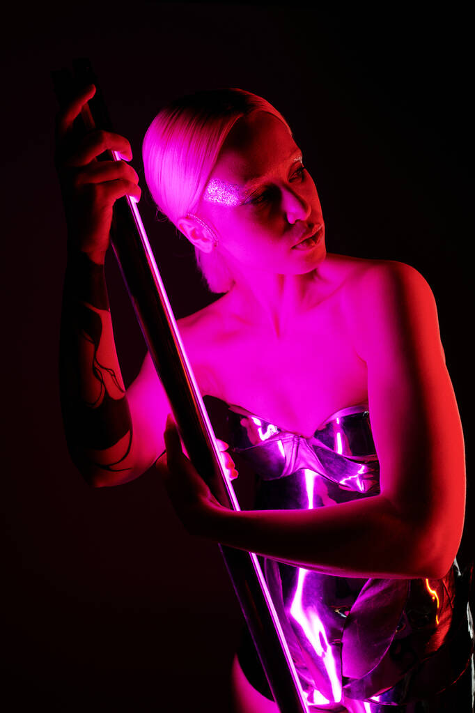 attraktive sonderbare Frau in futuristischer metallischer Kleidung, die einen rosafarbenen LED-Lampenstock hält und wegschaut - Foto, Bild