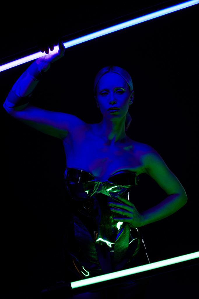 atractiva mujer extraña en traje robótico posando con palo de lámpara led azul y mirando a la cámara - Foto, Imagen