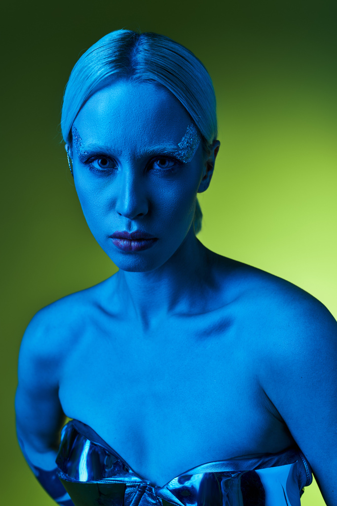 mujer extravagante en traje robótico posando en luces azules sobre fondo verde y mirando a la cámara - Foto, Imagen