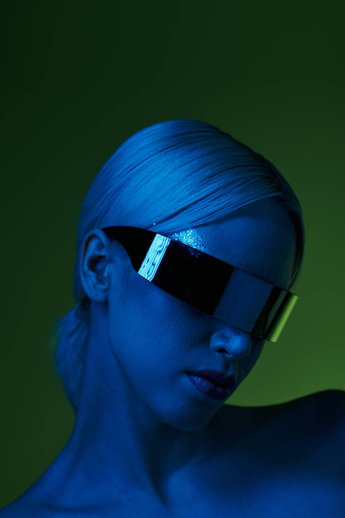 Bilim kurgu robotik güneş gözlüklü müsrif sarışın kadın yeşil arka planda mavi ışıkta poz veriyor. - Fotoğraf, Görsel