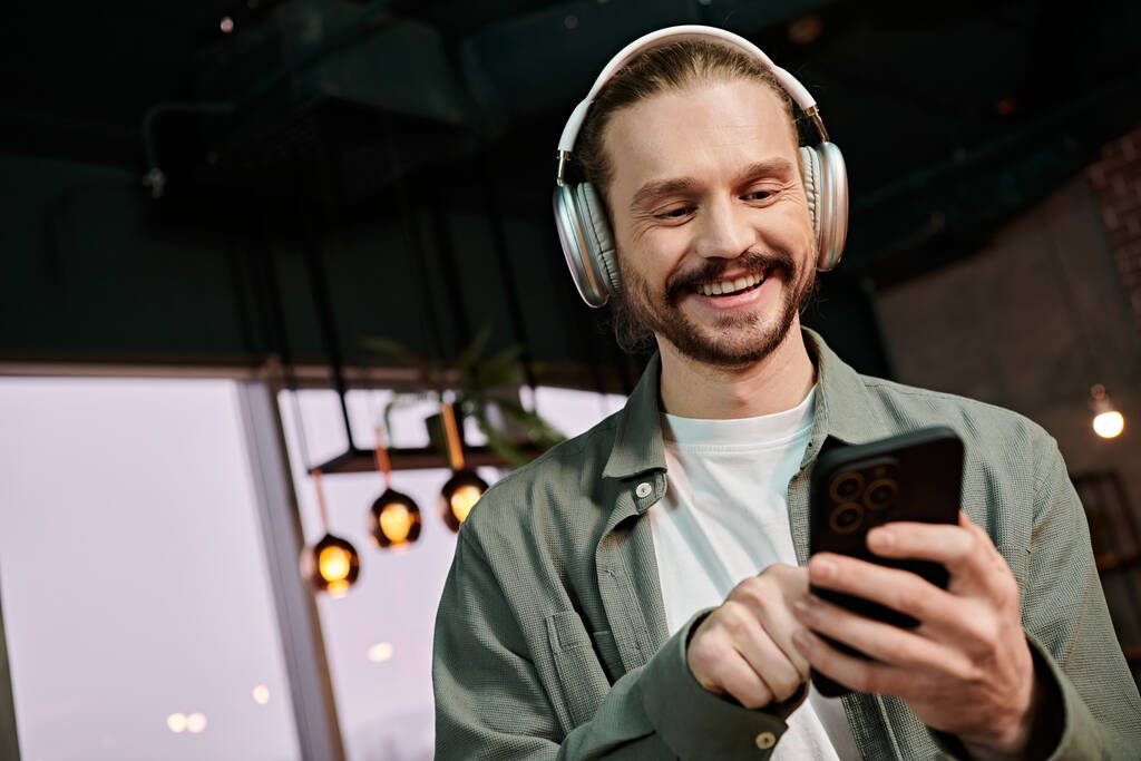 Een man met een koptelefoon op, met een mobiele telefoon, verdwaald in muziek en conversatie in een modern café.. - Foto, afbeelding