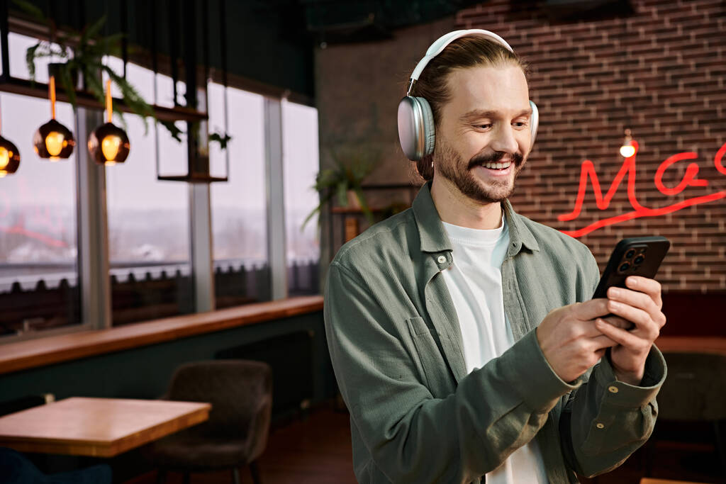 Mężczyzna, noszący słuchawki, gapiący się na telefon w nowoczesnej kawiarni, zanurzony we własnym świecie muzyki.. - Zdjęcie, obraz