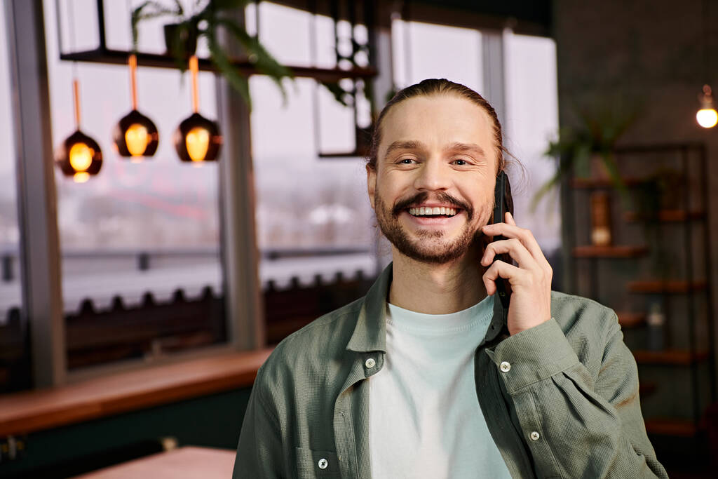 Mann mit stylischem Bart unterhält sich lebhaft auf einem Handy in einem modernen Café-Ambiente. - Foto, Bild