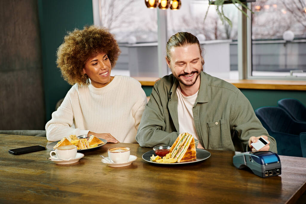 Una mujer afroamericana y un hombre sentado en una mesa, disfrutando de platos de comida en un moderno café. - Foto, imagen