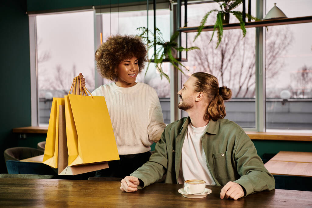 Μια κομψή Αφρο-Αμερικανίδα και ένας άντρας με τσάντες για ψώνια, κάθονται σε ένα τραπέζι σε ένα μοντέρνο καφέ. - Φωτογραφία, εικόνα