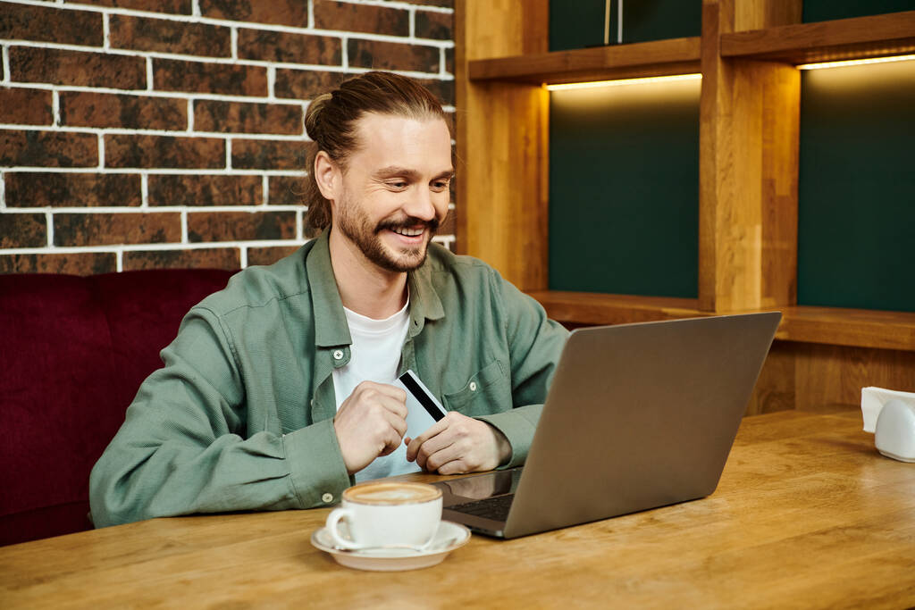 Ένας άνδρας κάθεται σε ένα σύγχρονο καφέ, επικεντρωμένος στην οθόνη του υπολογιστή του, που ασχολείται με δραστηριότητες εργασίας ή αναψυχής. - Φωτογραφία, εικόνα