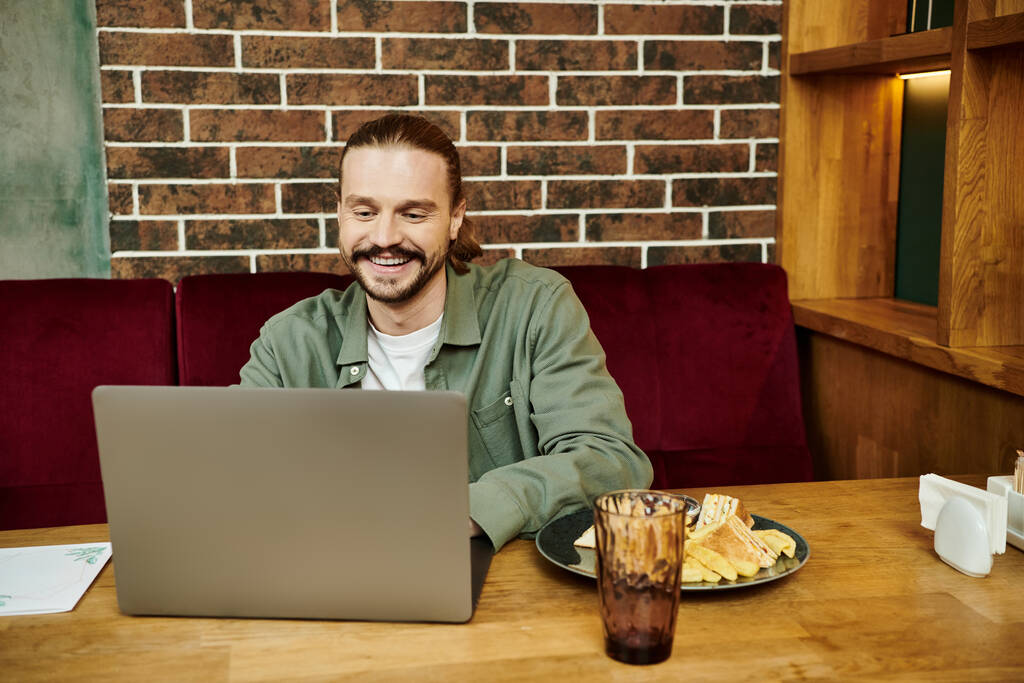 Bir adam, modern bir kafe ortamında dizüstü bilgisayarına odaklanmış bir şekilde masada oturuyor.. - Fotoğraf, Görsel