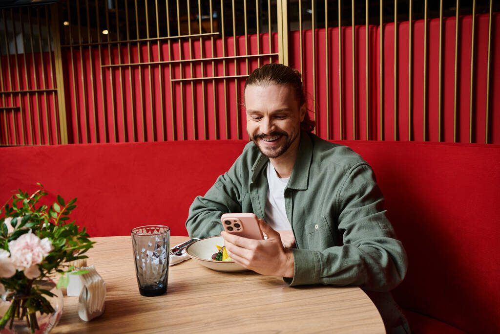 Mies istuu pöydän ääressä, uppoutuneena kännykkäänsä modernissa kahvilassa. - Valokuva, kuva