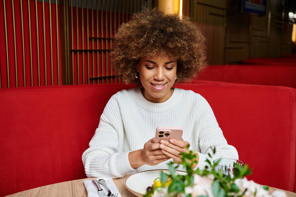 Стильная афроамериканка погружается в свой телефон, сидя за столиком в кафе. - Фото, изображение