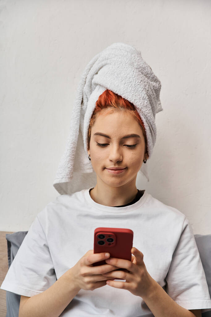 νεαρός υπερβολικός άνθρωπος στο homewear με πετσέτα μαλλιών κοιτάζοντας το smartphone της, ενώ χαλαρώνοντας στο κρεβάτι - Φωτογραφία, εικόνα