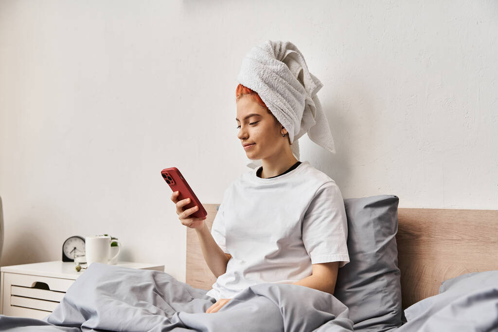 jong extravagant persoon in huiskleding met haardoek kijken naar haar smartphone terwijl ontspannen in bed - Foto, afbeelding